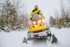 A man rides a yellow snowmobile through a winter forest